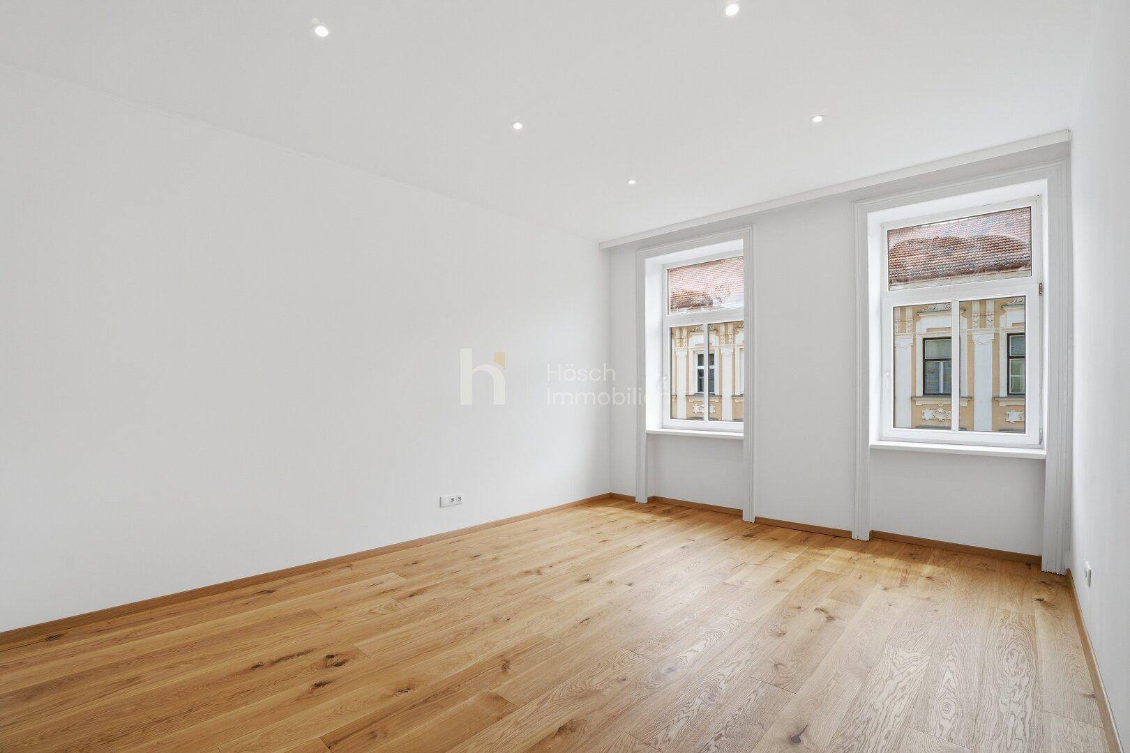 Heller, leerer Raum mit Holzboden und zwei Fenstern, ideal für verschiedene Nutzungsmöglichkeiten.