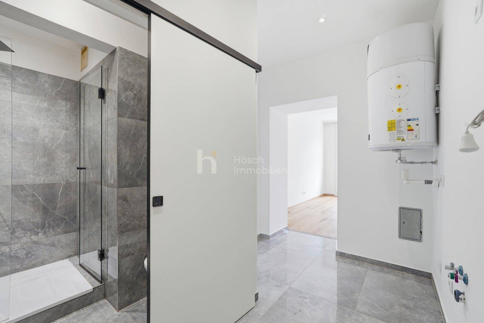 Modernes Badezimmer mit begehbarer Dusche, grauen Fliesen und einem Warmwasserboiler an der Wand.
