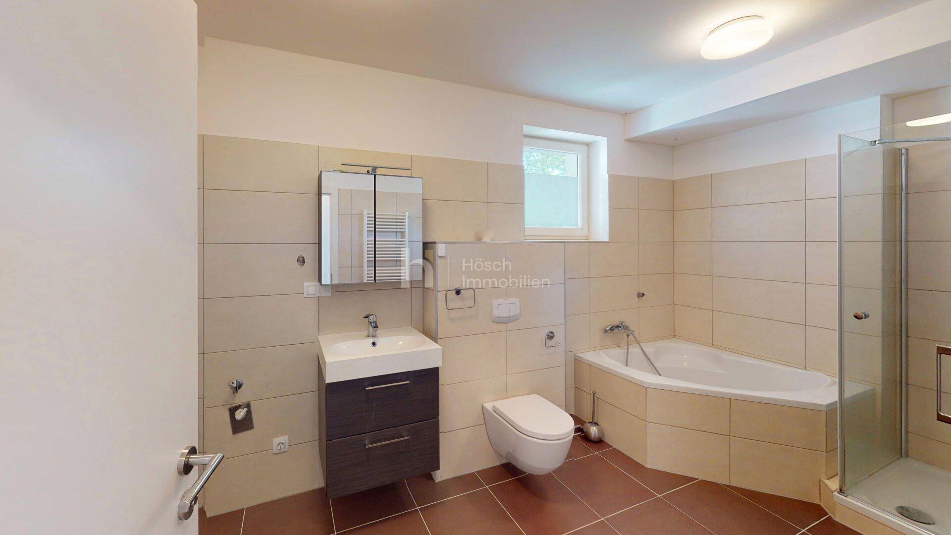 Badezimmer mit beigefarbenen Fliesen, Eckbadewanne, Dusche und Fenster.