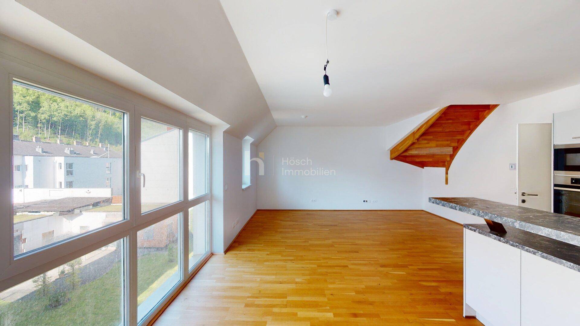 Geräumiger Wohnbereich mit Holzboden, großen Fenstern und einer offenen Holztreppe.