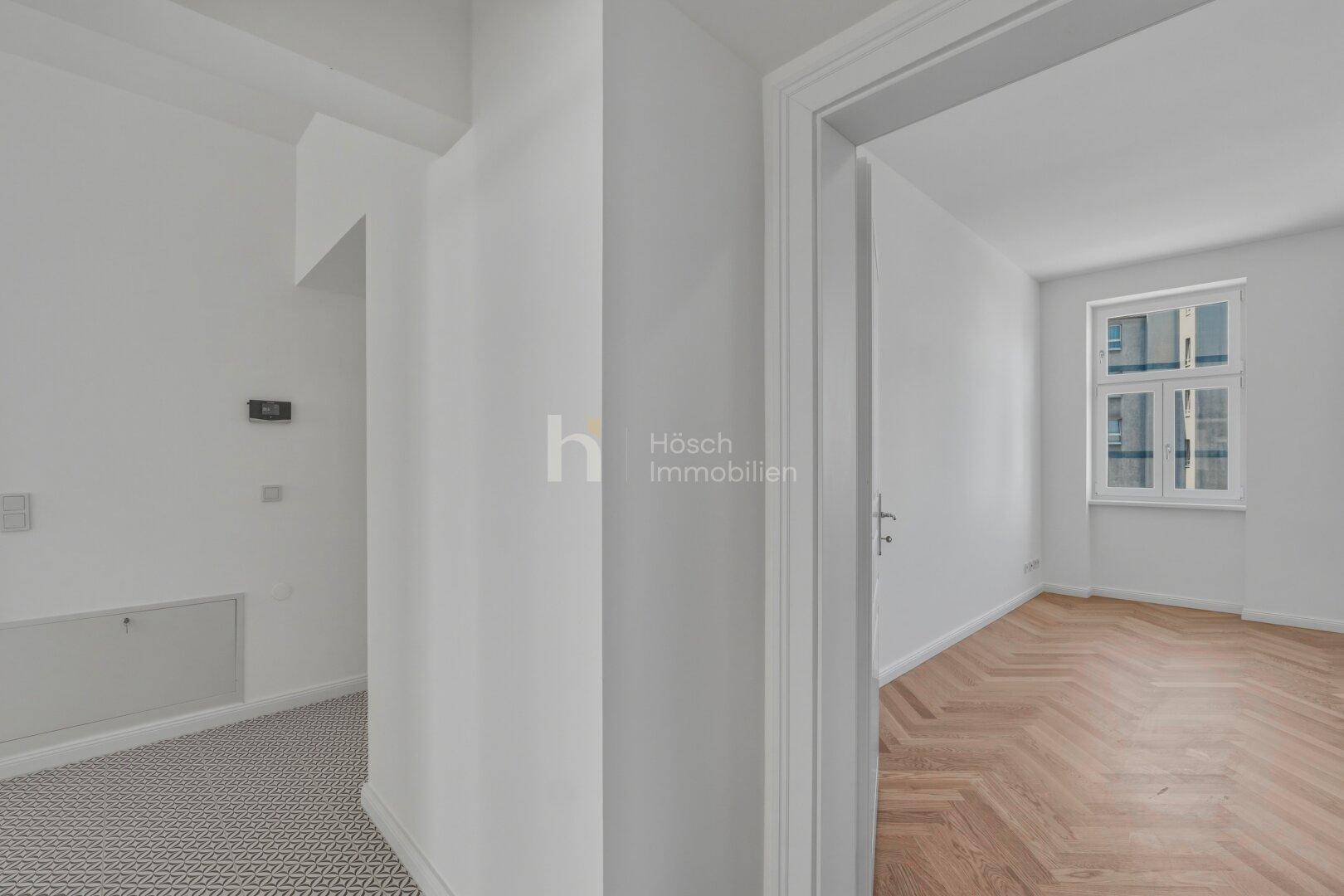 Heller Flur mit modernem Bodenbelag und Blick in ein Zimmer mit Fischgrätparkett und großen Fenstern.