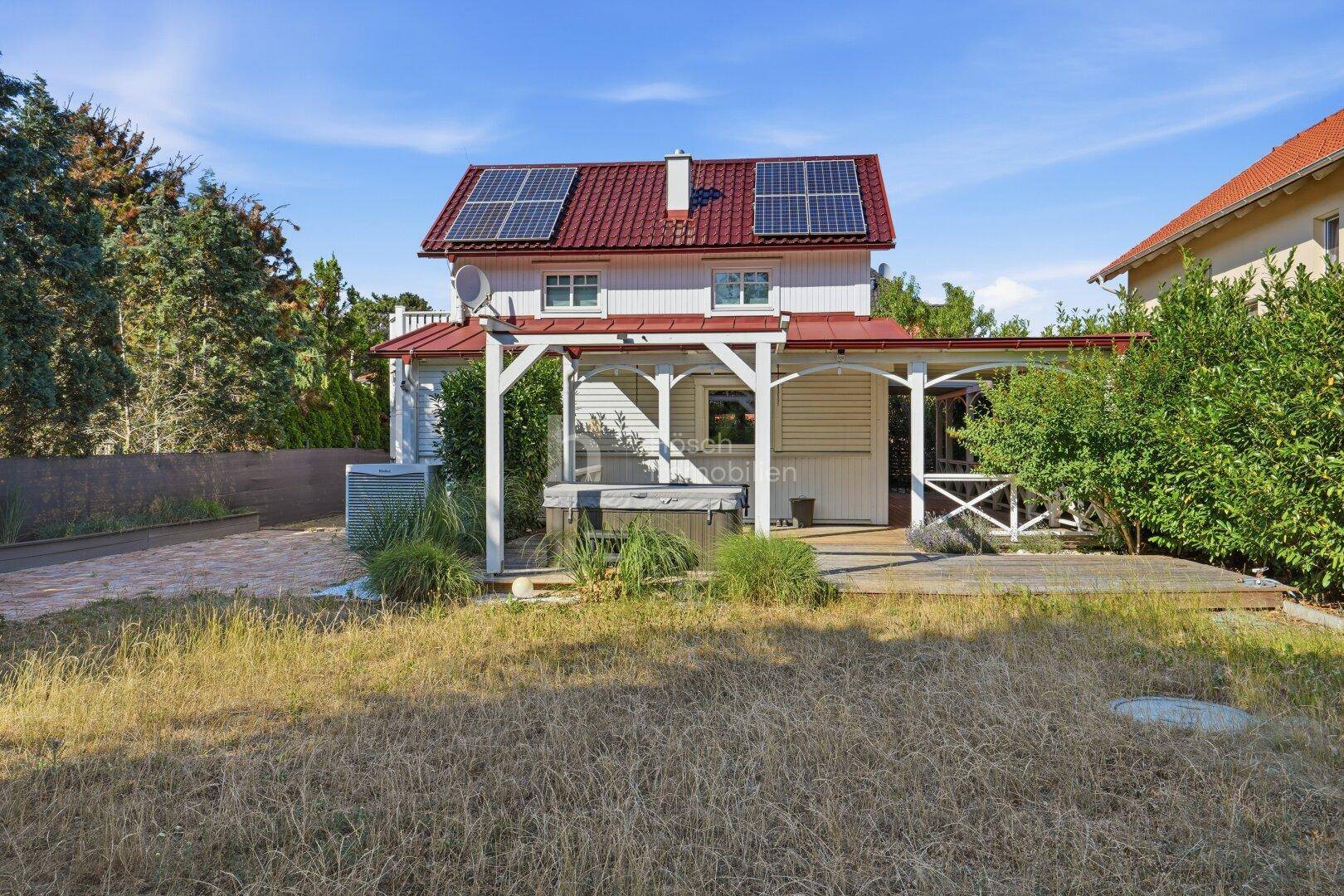 Großer Gartenbereich mit Rasenfläche, Holzterrasse, Whirlpool und einem Haus mit Solaranlage auf dem Dach.