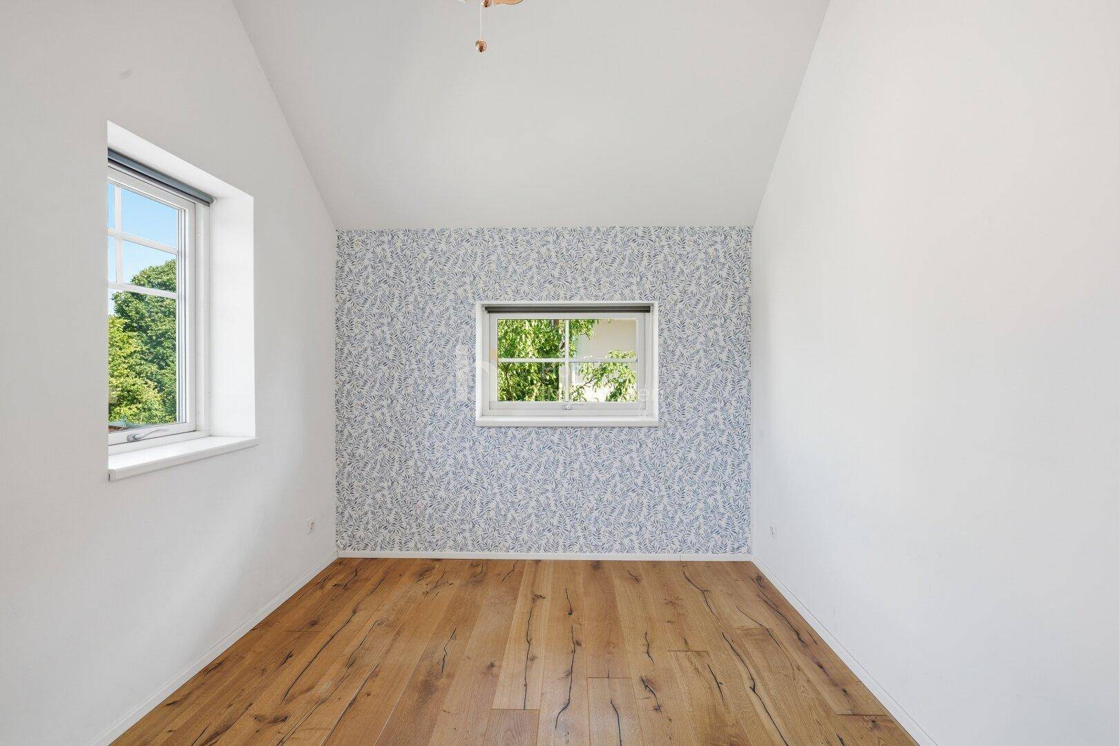 Gemütliches Schlafzimmer mit Holzboden, einer gemusterten Tapetenwand und einem Fenster mit Blick ins Grüne.