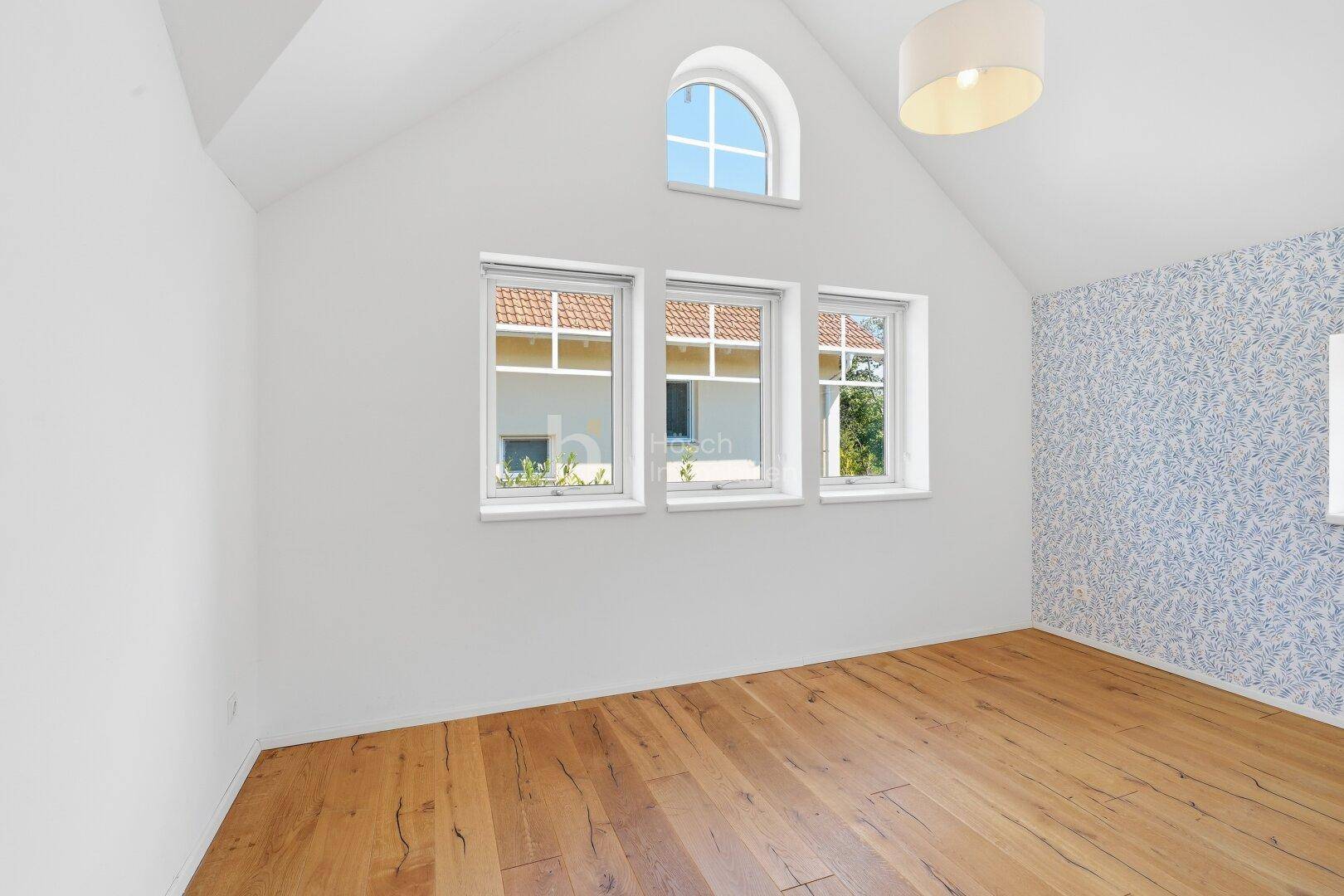 Schlafzimmer mit Holzboden, Dachschräge und drei Fenstern, die viel Tageslicht hereinlassen.
