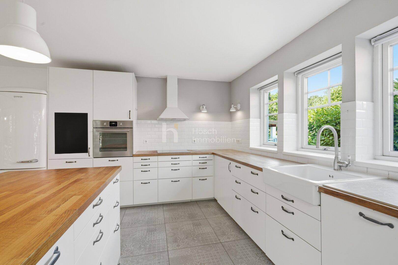 Geräumige weiße Küche mit Holz-Arbeitsplatten, integriertem Backofen und einem großen Spülbecken unter den Fenstern.