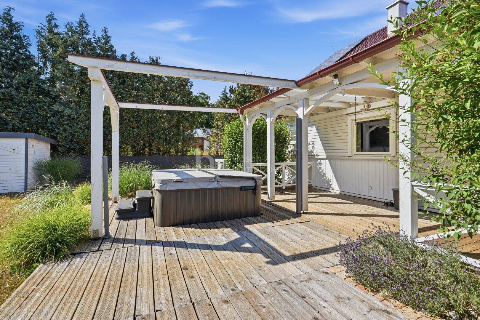 Holzterrasse mit einem Whirlpool unter einer Pergola, umgeben von Grünflächen und einem Gartenhaus.