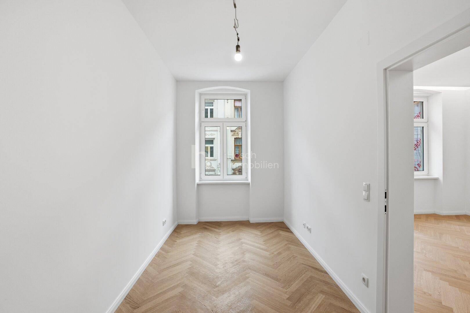Helles, schmales Zimmer mit Fischgrätparkett, weiß gestrichenen Wänden und einem Fenster.