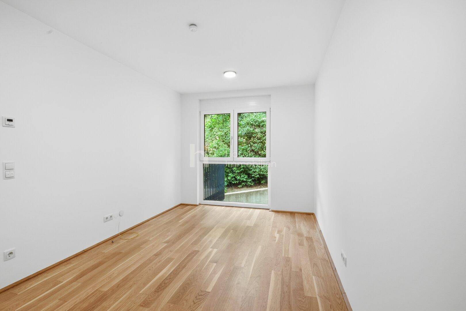 Helles Zimmer mit Holzboden und Fenster, das einen Blick ins Grüne bietet.