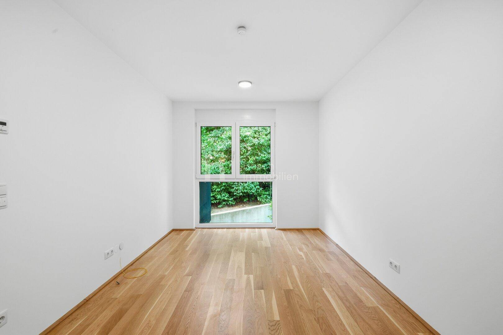 Helles Zimmer mit Holzboden und Fenster, das einen Blick ins Grüne bietet.