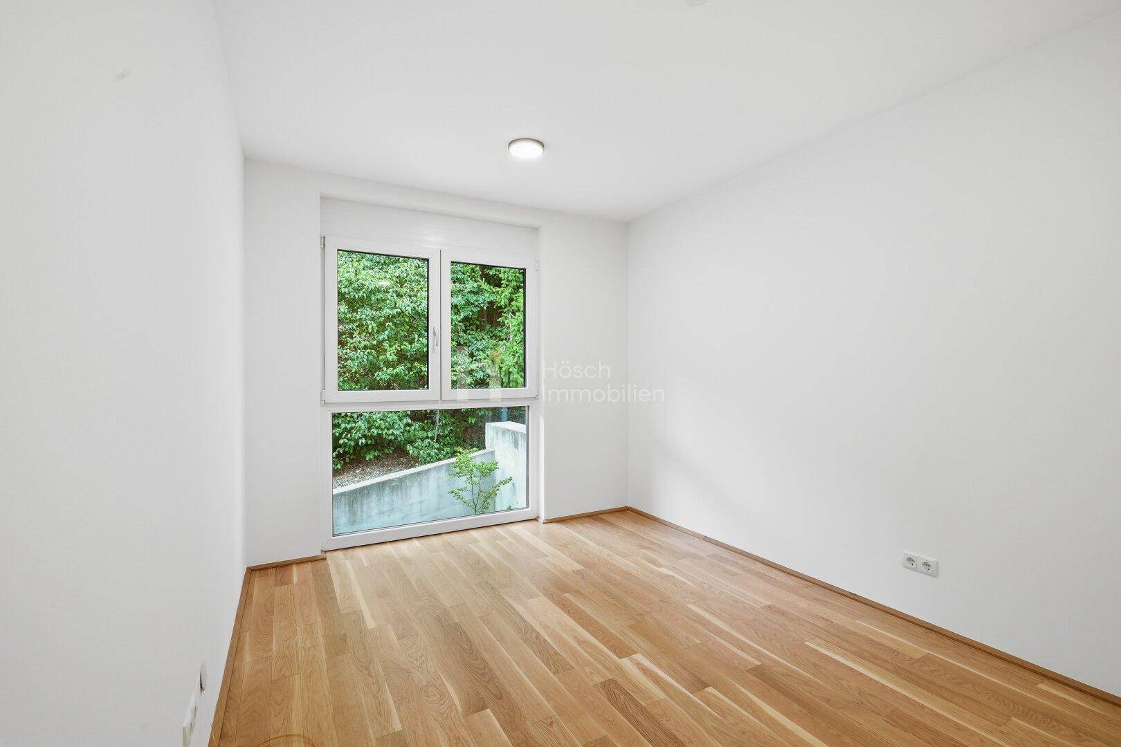 Helles Zimmer mit Holzboden und großem Fenster, das einen Blick ins Grüne bietet.
