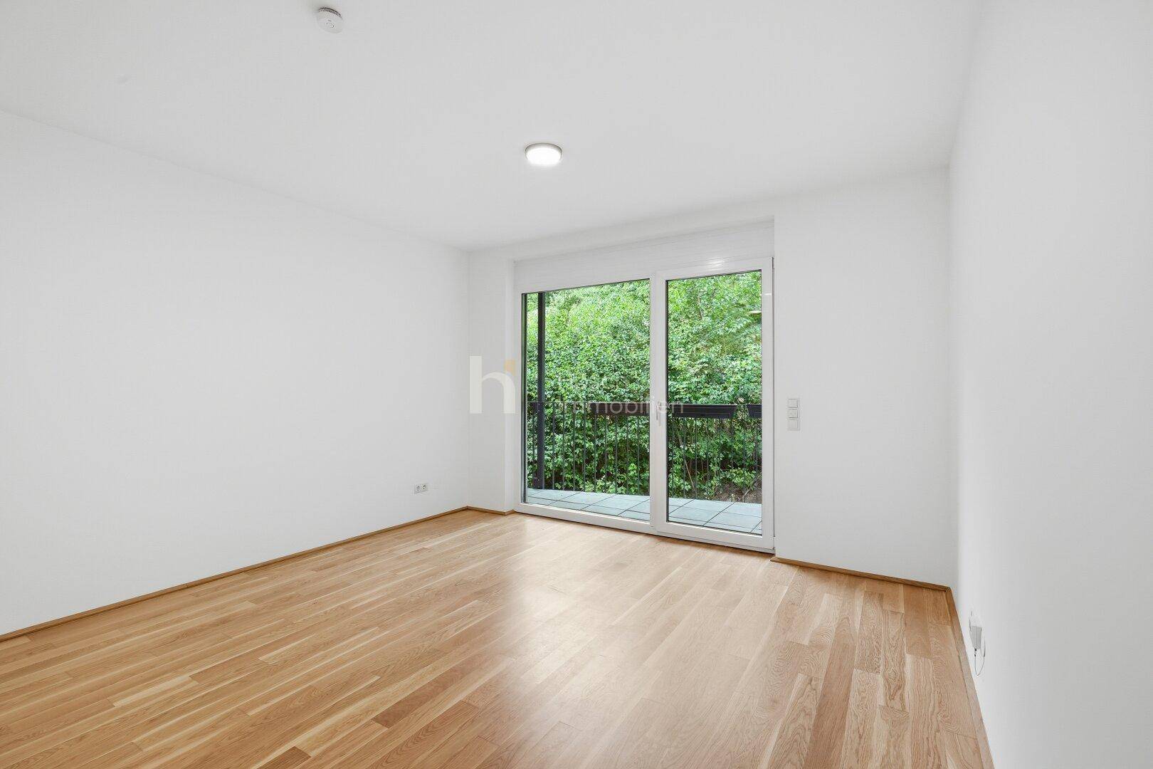 Helles Zimmer mit Holzboden und Zugang zu einem Balkon mit Blick ins Grüne.