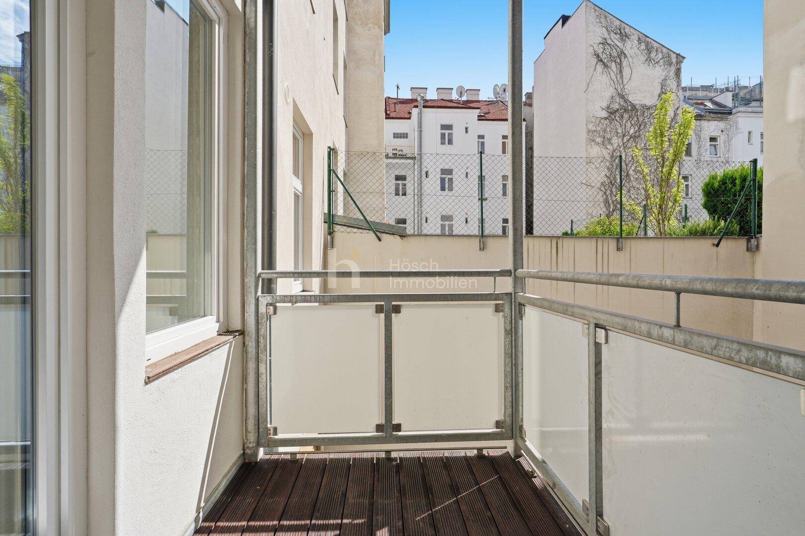 Schmaler Balkon mit Holzboden und Blick auf die umliegenden Gebäude und den Himmel.