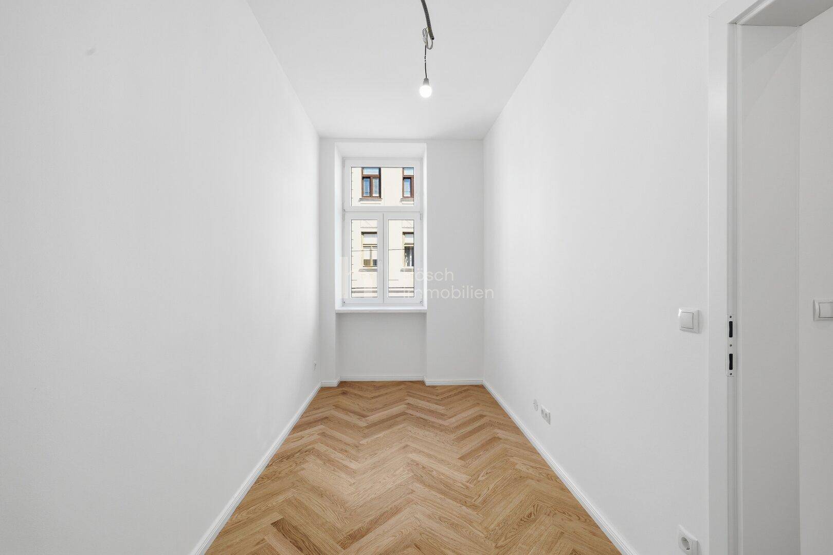 Schmaler Raum mit Fischgrätparkett und einem Fenster, das viel Tageslicht hereinlässt.