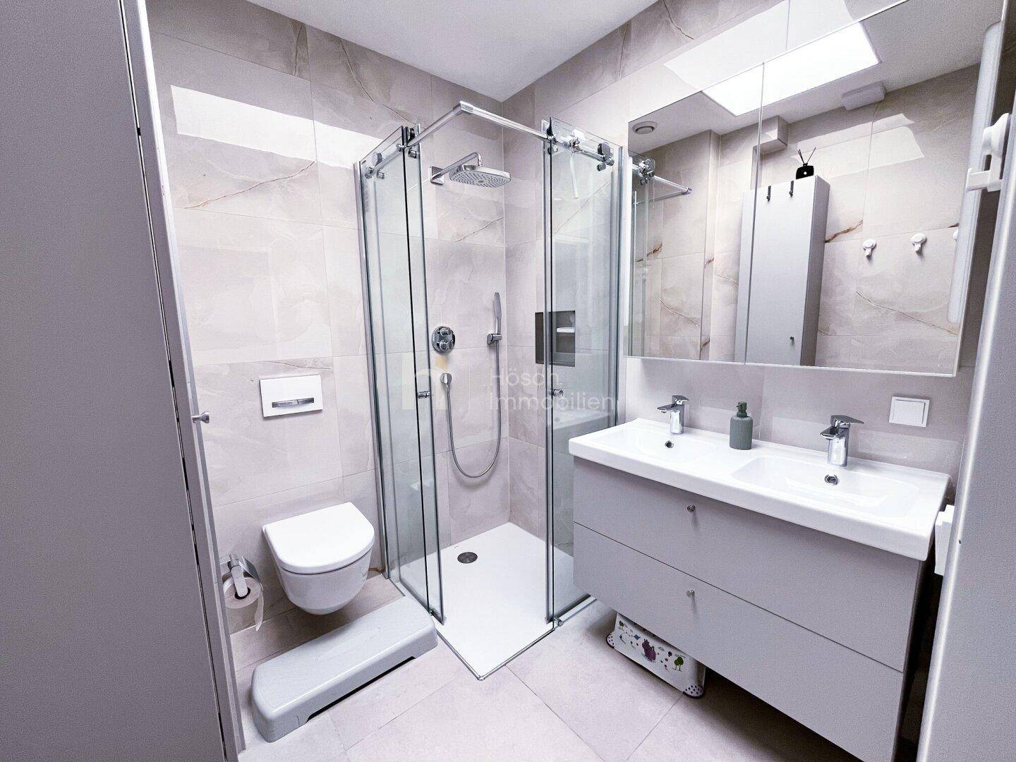 Modernes Badezimmer mit Doppelwaschbecken, wandhängender Toilette und geräumiger Glasdusche.
