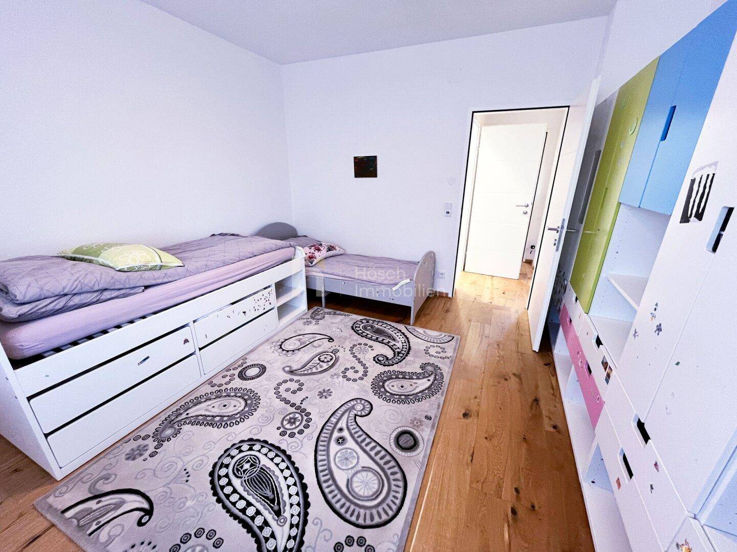Kinderzimmer mit zwei Betten, farbenfrohem Teppich und einem bunten Kleiderschrank.