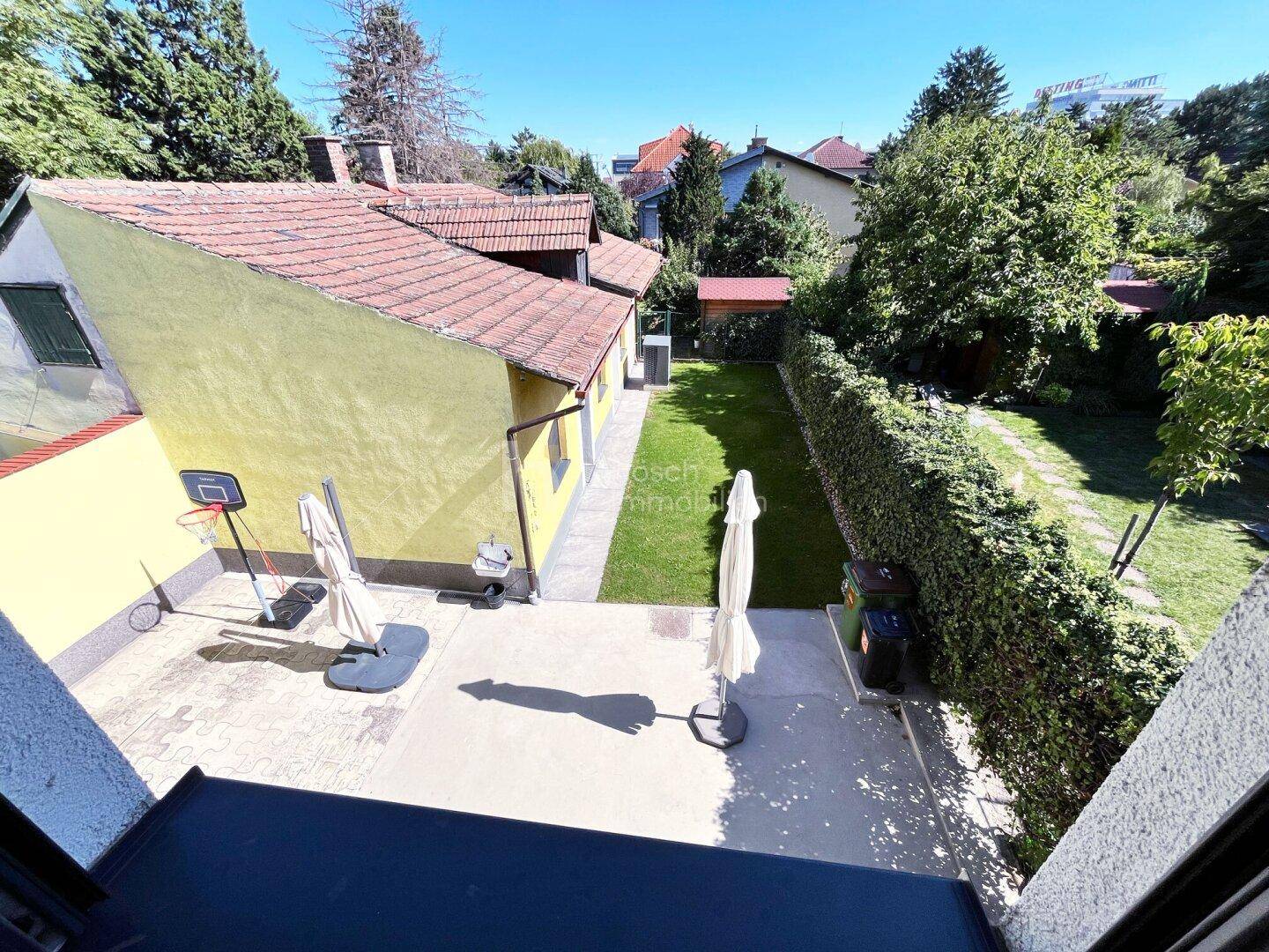 Blick von oben auf den gepflegten Garten mit Terrasse, Rasenfläche und umgebendem Grün.