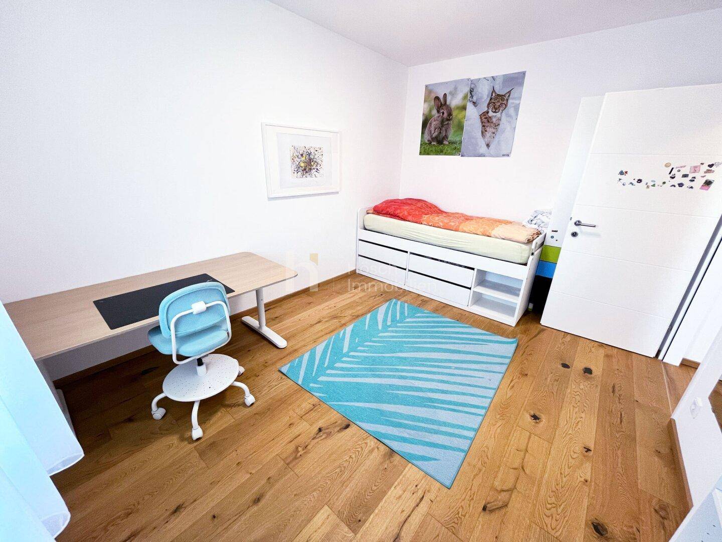 Helles Kinderzimmer mit Schreibtisch, Bett und einem farbenfrohen Teppich auf dem Holzboden.
