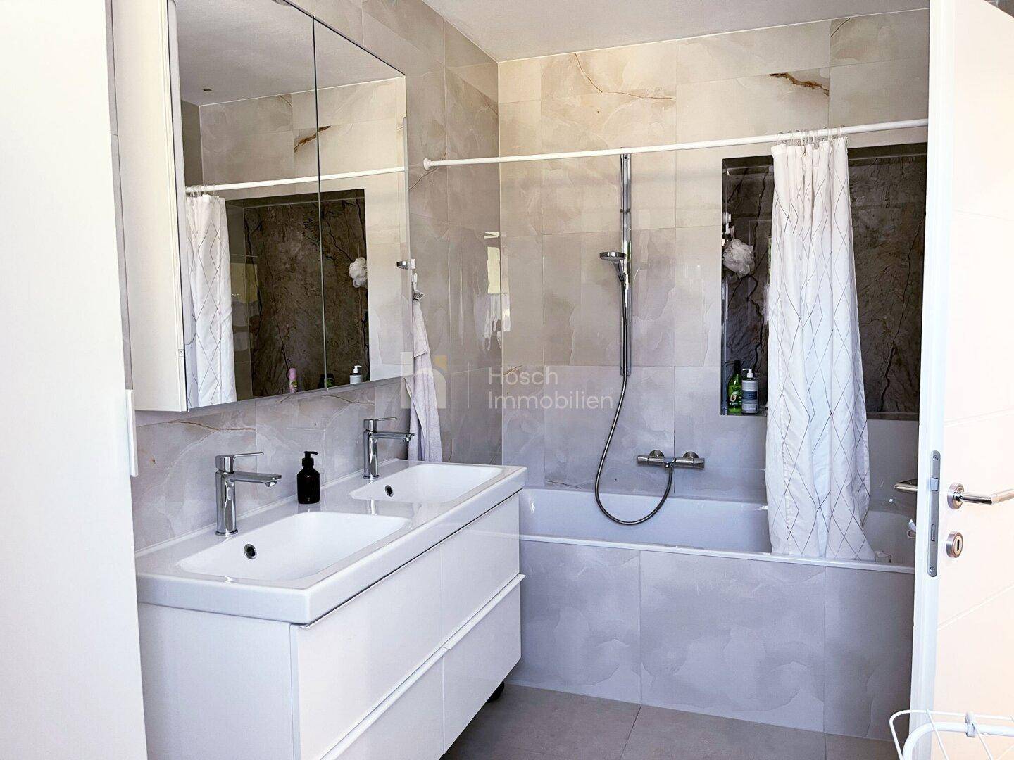 Geräumiges Badezimmer mit Doppelwaschbecken, Badewanne und Duschvorhang, hell gefliest.