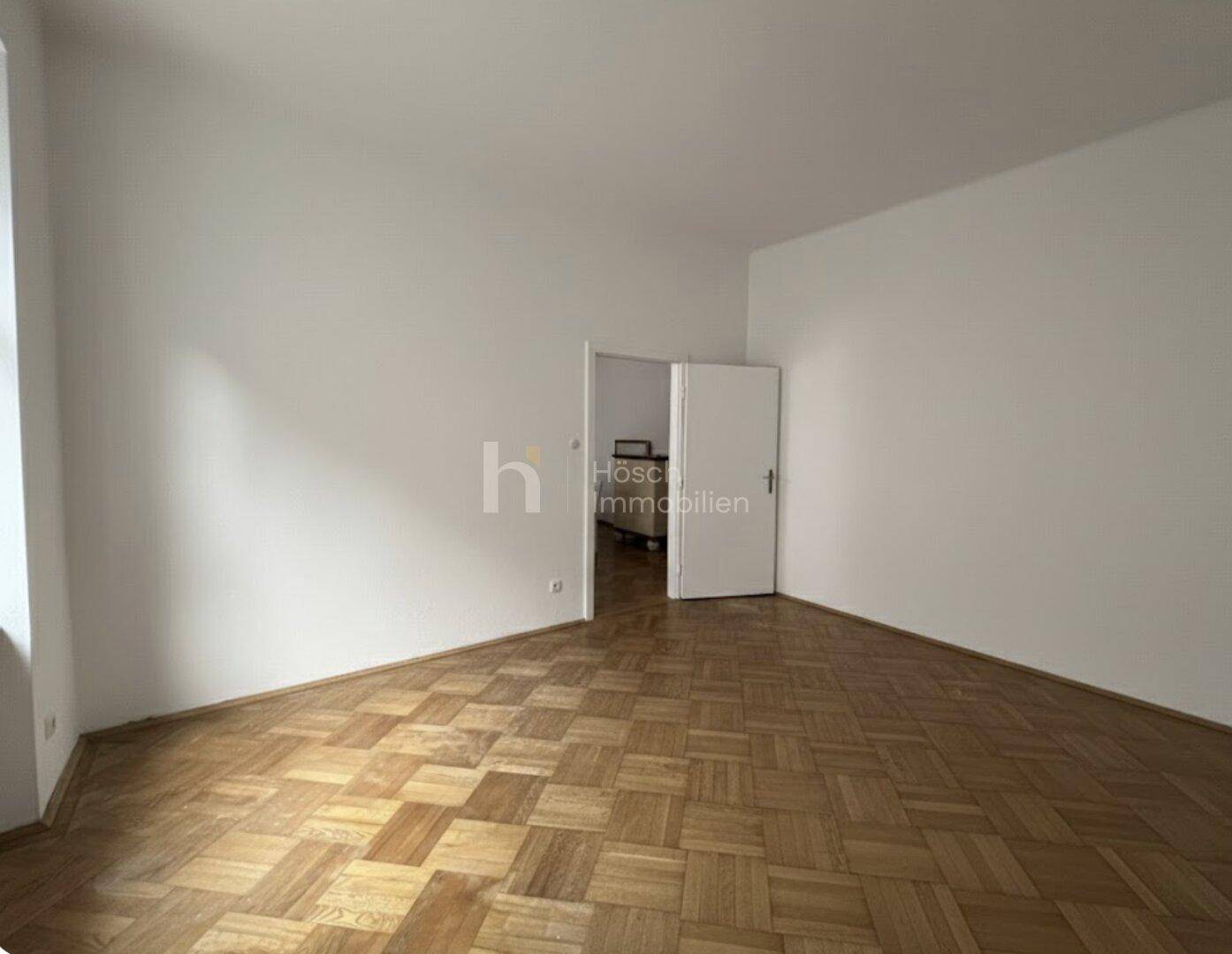 Unmöbliertes Zimmer mit Fischgrätparkett und einer Tür, die zu einem weiteren Bereich führt.