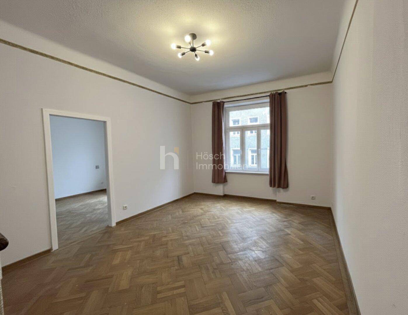 Leeres Zimmer mit Fischgrätparkett, einem Fenster und Zugang zu einem weiteren Raum.