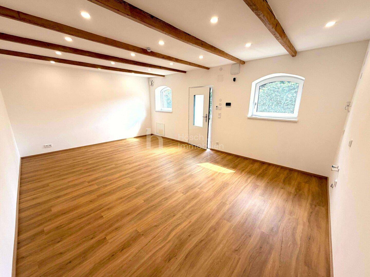 Heller, leerer Raum mit sichtbaren Holzbalken an der Decke und Laminatboden.