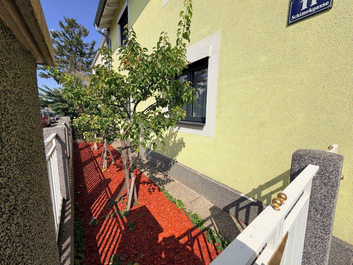 Seitenansicht des Hauses mit einem schmalen Gartenstreifen und jungen Bäumen unter blauem Himmel.