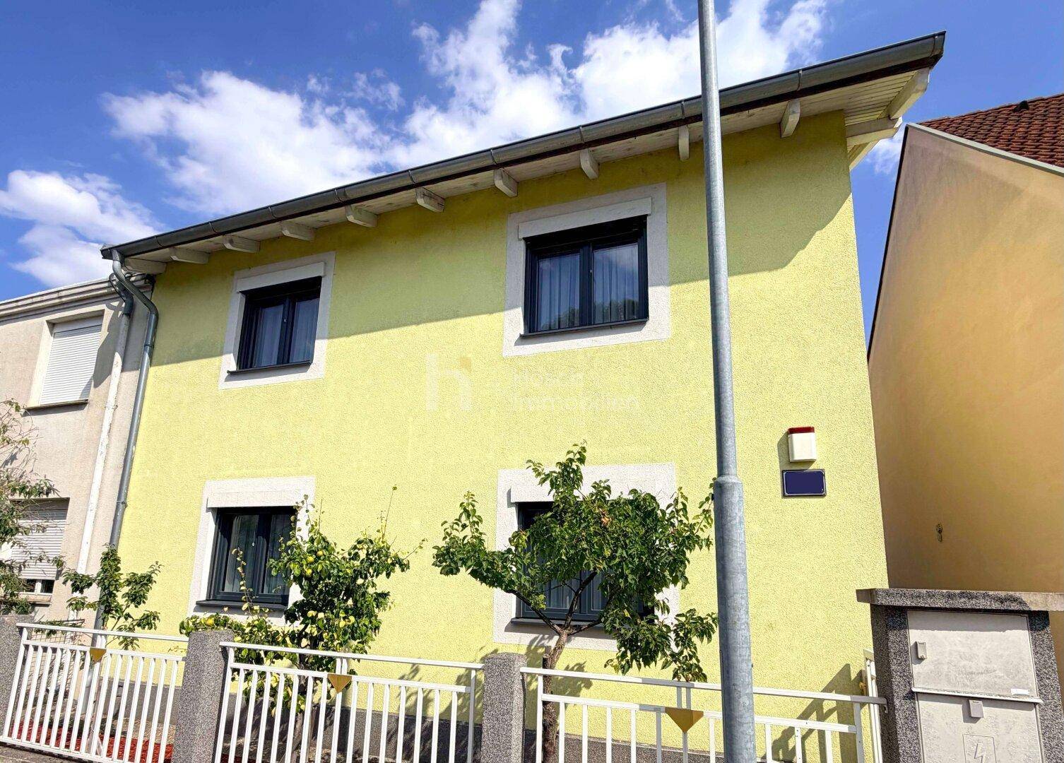 Gelbe Fassade eines Einfamilienhauses mit dunklen Fenstern und weißem Zaun unter blauem Himmel.