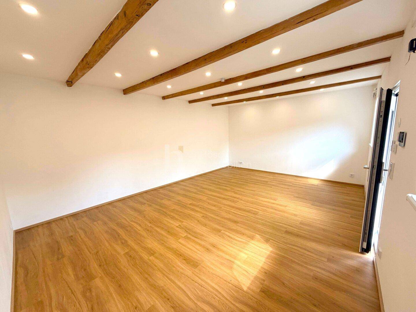 Geräumiger, unmöblierter Raum mit sichtbaren Holzbalken und Einbaustrahlern an der Decke.