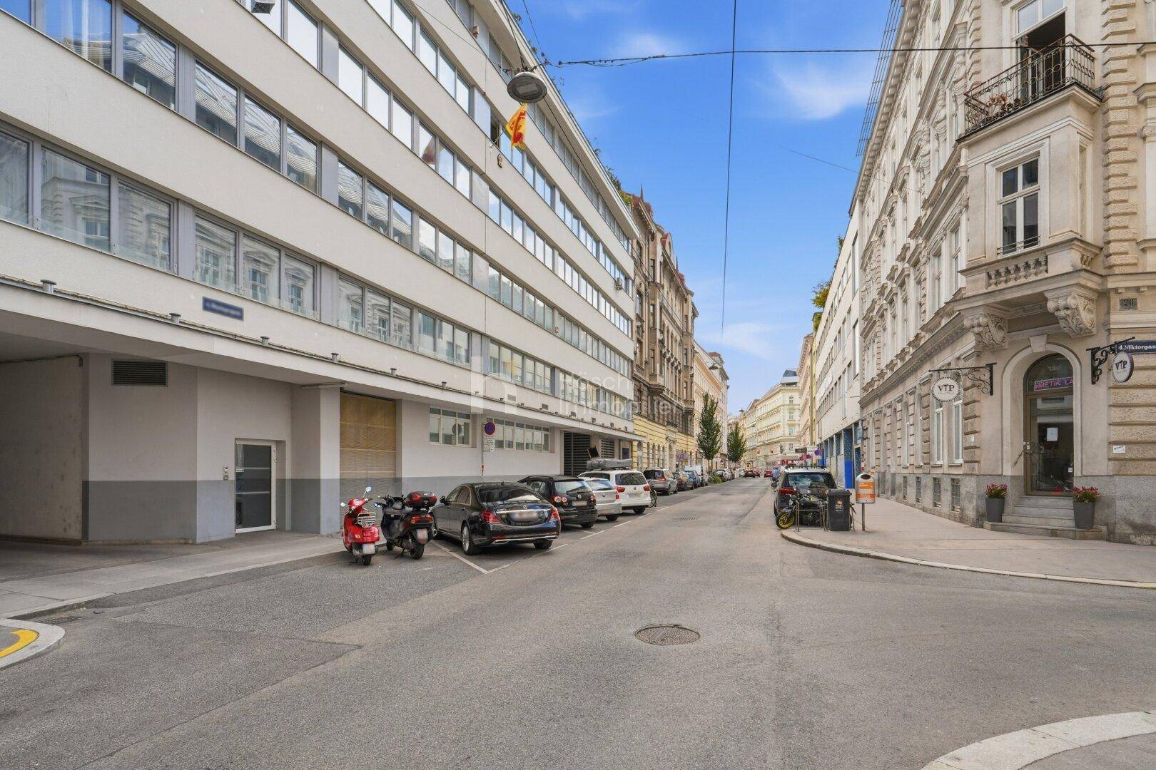 Städtische Straße mit moderner Gebäudefassade, parkenden Autos und klassischer Architektur auf der rechten Seite.