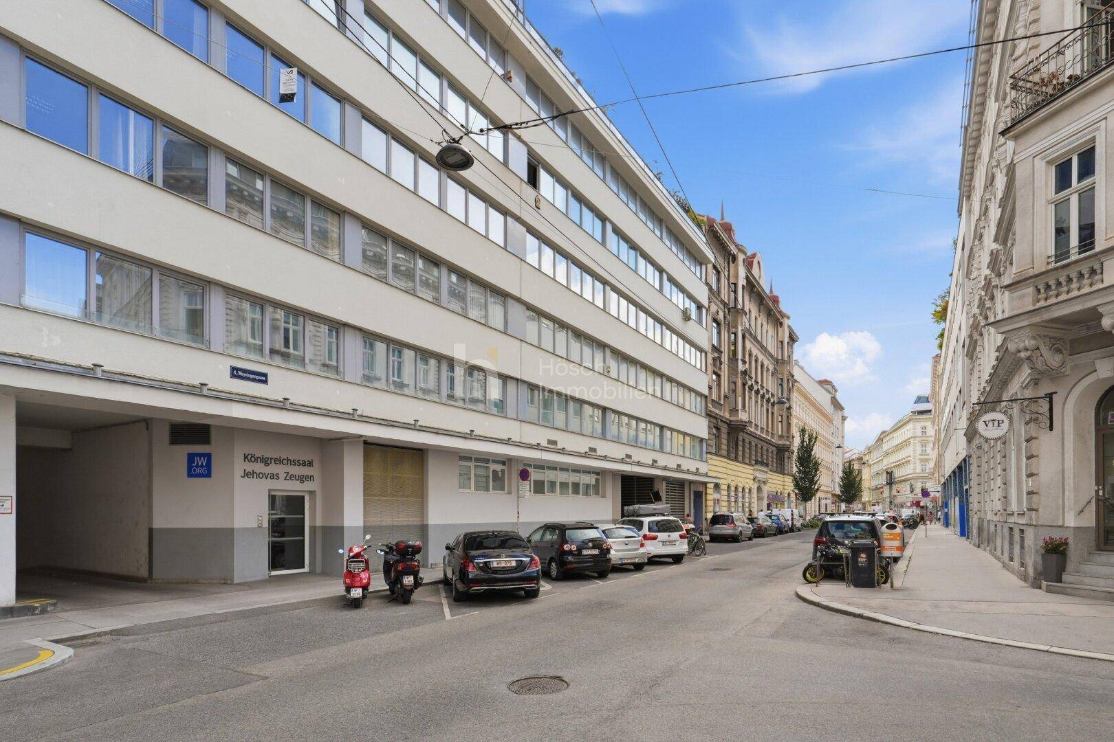 Städtische Straße mit moderner Architektur und traditionellen Gebäuden, gesäumt von parkenden Fahrzeugen.