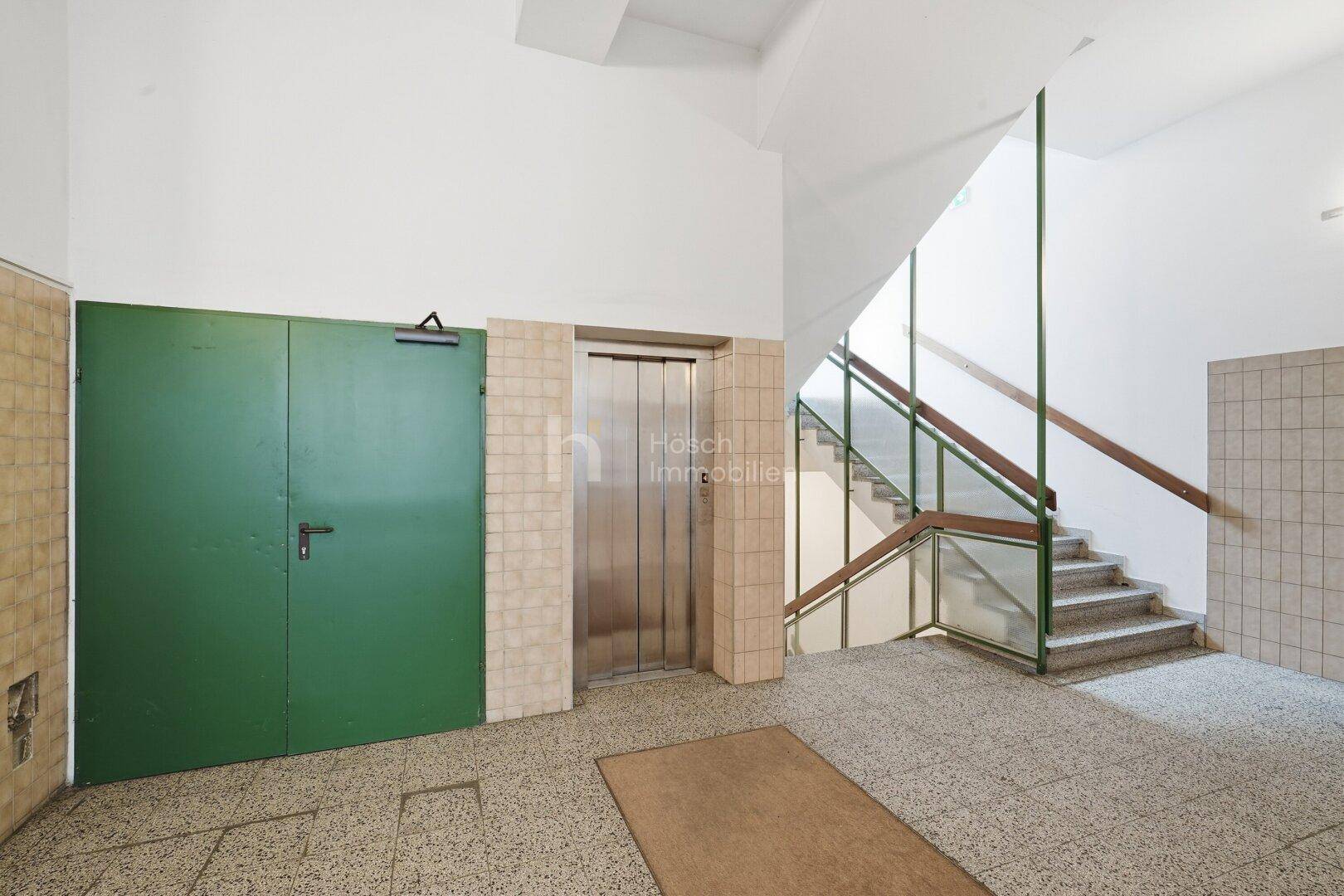 Eingangsbereich mit Aufzug, Treppenhaus und Fliesenboden in einem gut erhaltenen älteren Gebäude.
