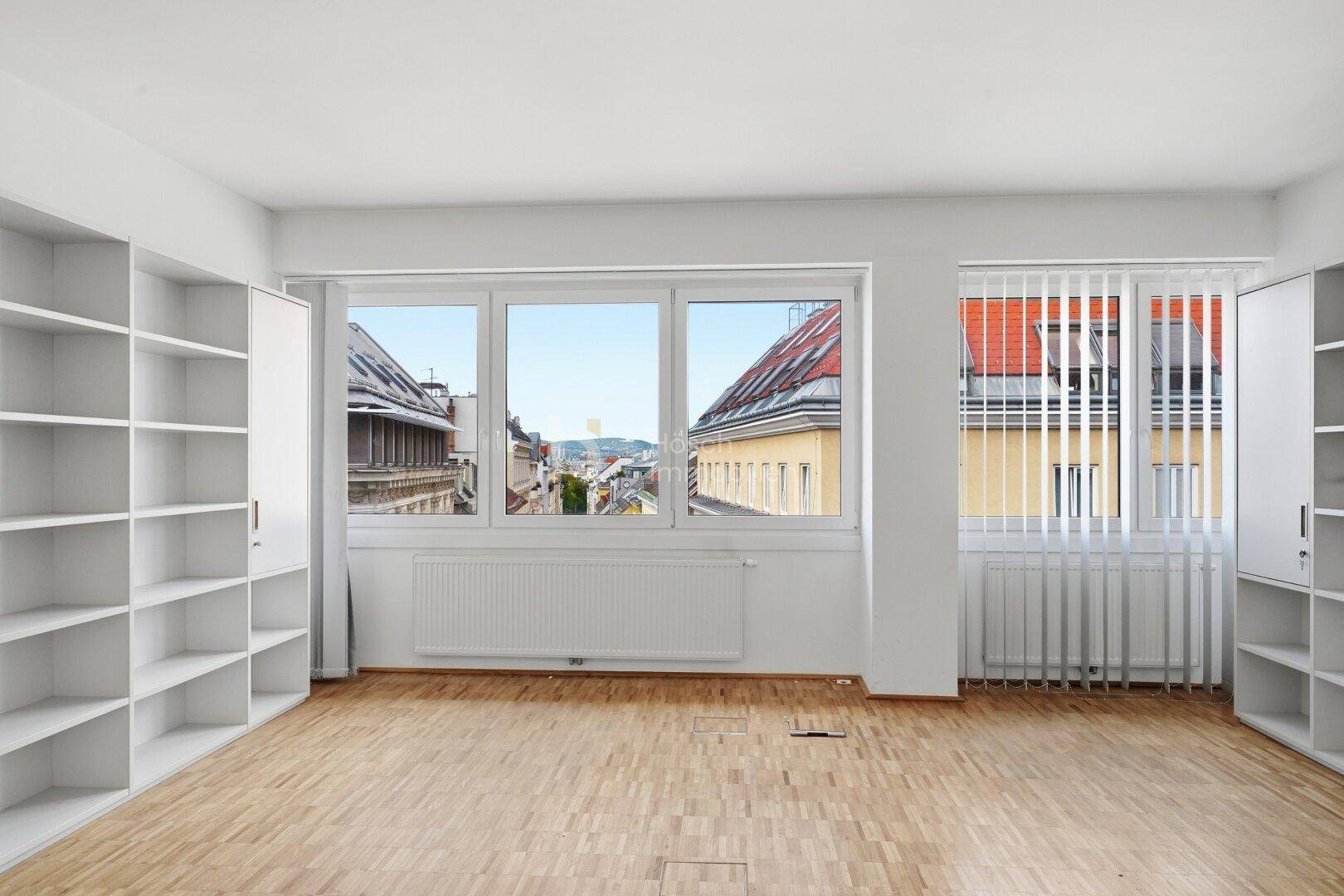 Heller Büroraum mit Parkettboden, weißen Regalen und Blick auf die Dächer der Stadt.
