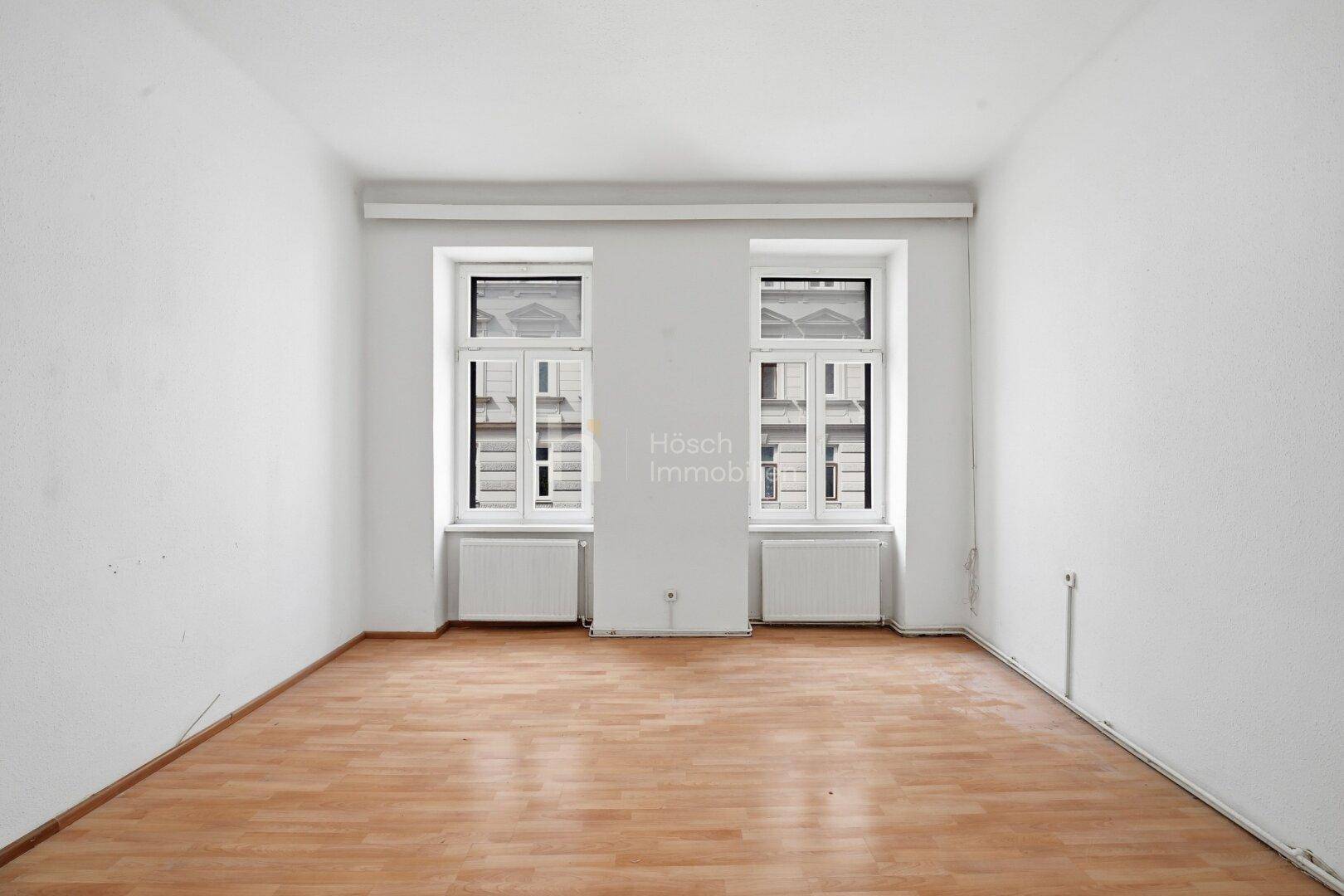 Leerer Raum mit zwei Fenstern, die auf eine Gebäudefassade blicken, und einem Laminatboden.