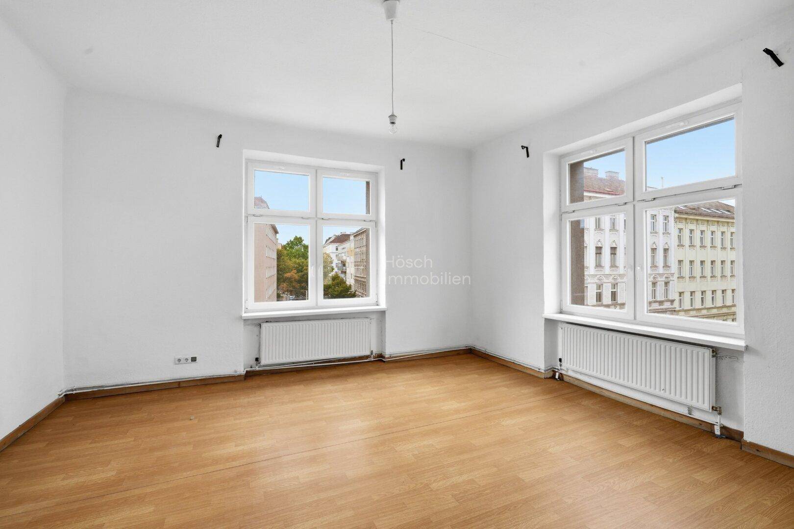 Leerer Raum mit Laminatboden und zwei Fenstern, die sowohl Grünflächen als auch Gebäude zeigen.