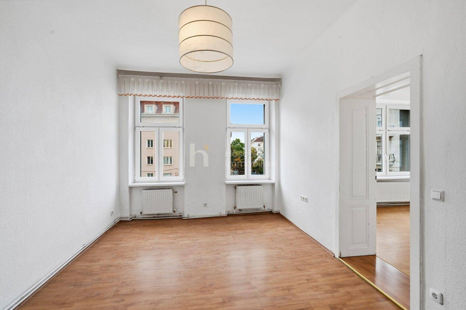Helles Zimmer mit Laminatboden und zwei Fenstern, die einen Blick auf die Stadt und Bäume ermöglichen.