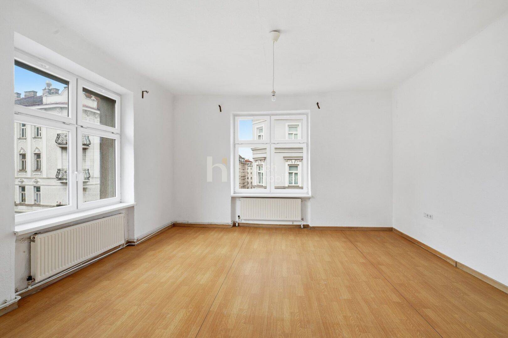 Geräumiges, leeres Zimmer mit Laminatboden und zwei großen Fenstern, die viel Tageslicht hereinlassen.