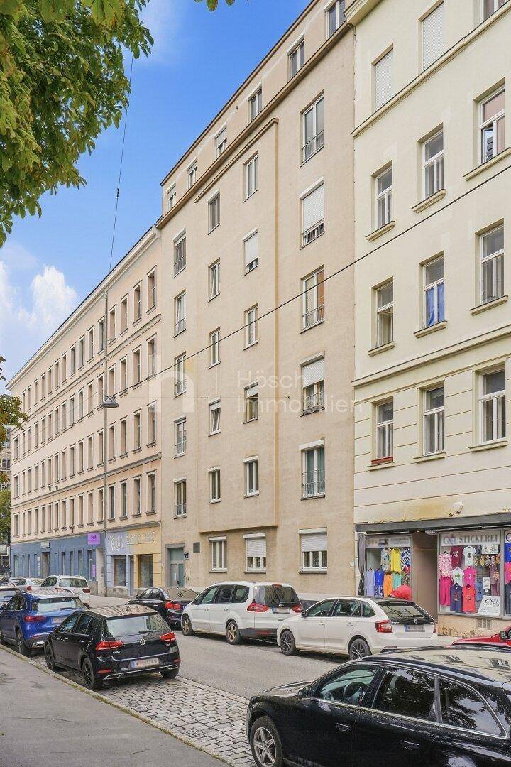 Mehrstöckiges Wohngebäude an einer Stadtstraße mit geparkten Autos und Geschäften im Erdgeschoss.