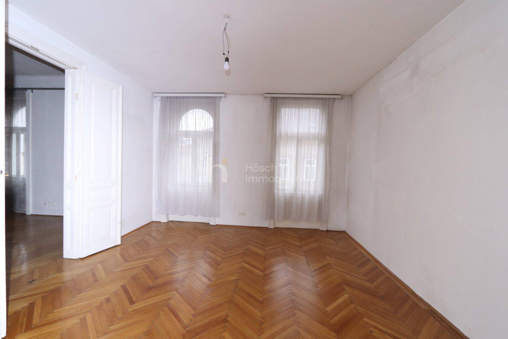 Geräumiges Zimmer mit klassischem Fischgrätparkett und zwei Fenstern, die viel Tageslicht hereinlassen.