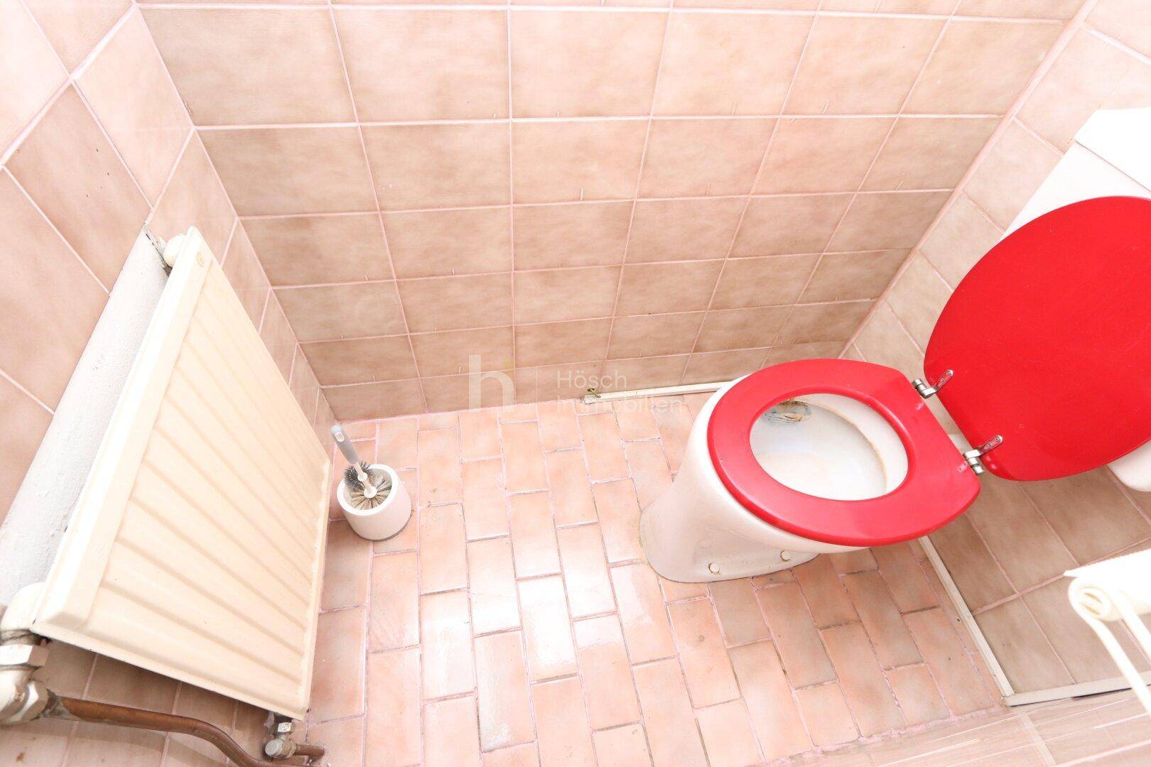 Kleines WC mit beigefarbenen Fliesen, Toilette mit rotem Sitz und einem Heizkörper.