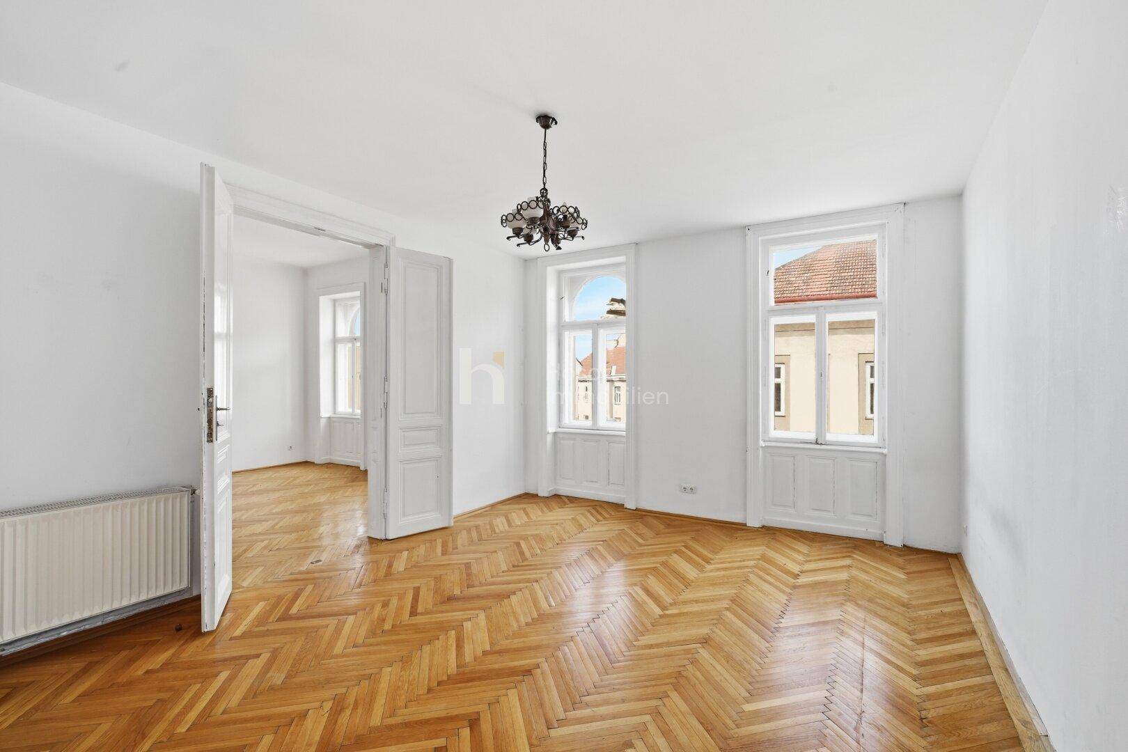 Geräumiges Zimmer mit Fischgrätparkett, hohen Decken und Blick auf die umliegenden Gebäude.