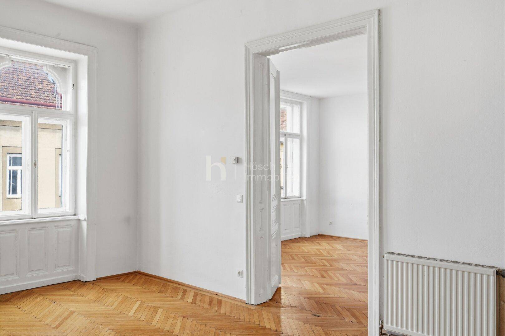 Heller Raum mit Parkettboden und Heizkörper, der durch eine offene Tür mit einem weiteren Zimmer verbunden ist.