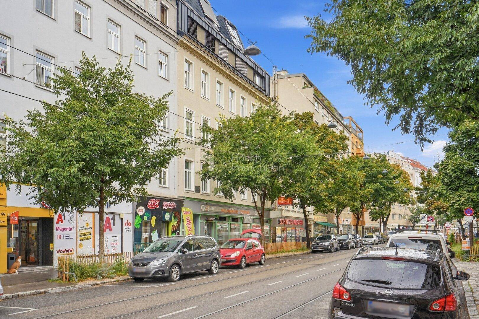 Ansicht eines Mehrfamilienhauses mit Geschäften im Erdgeschoss und parkenden Autos entlang der Straße.