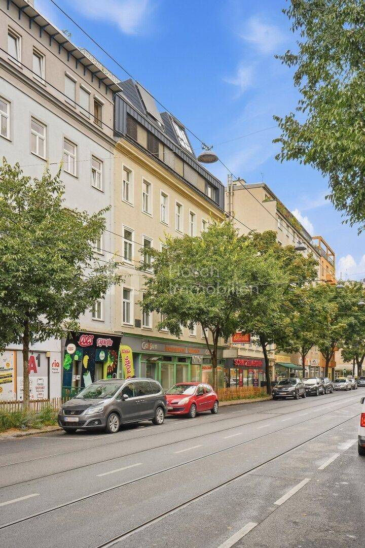 Belebtes Stadtbild mit einem Mehrfamilienhaus, Geschäften im Erdgeschoss und parkenden Autos.