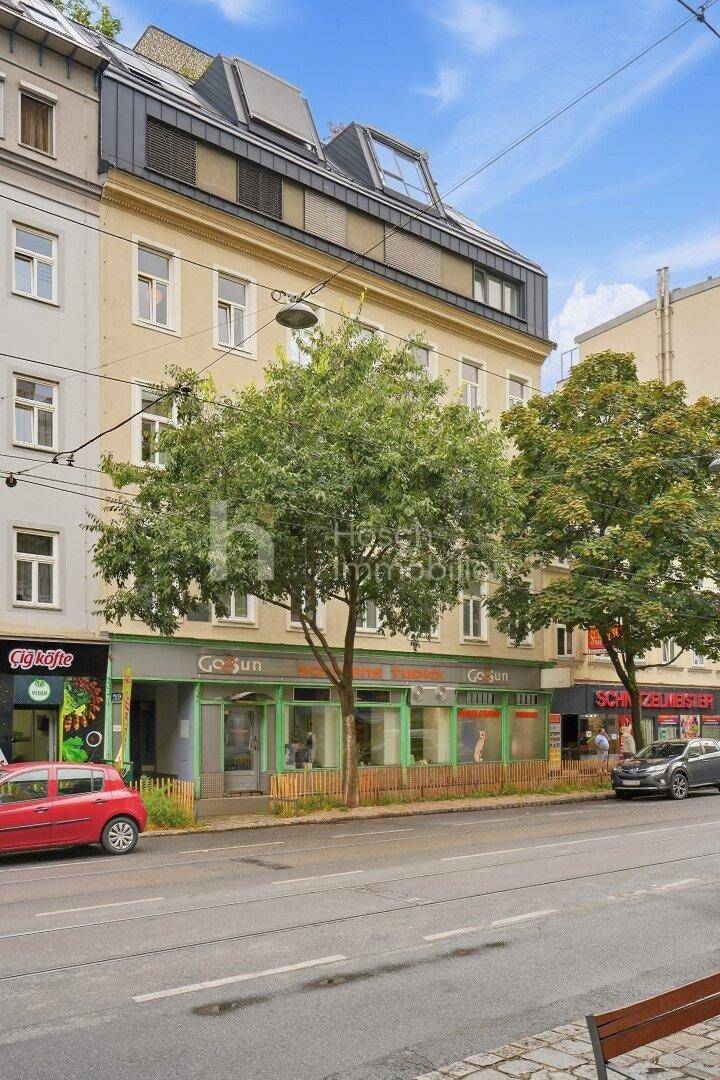 Fassade eines Wohngebäudes mit Geschäften im Erdgeschoss und modernen Dachgauben unter blauem Himmel.