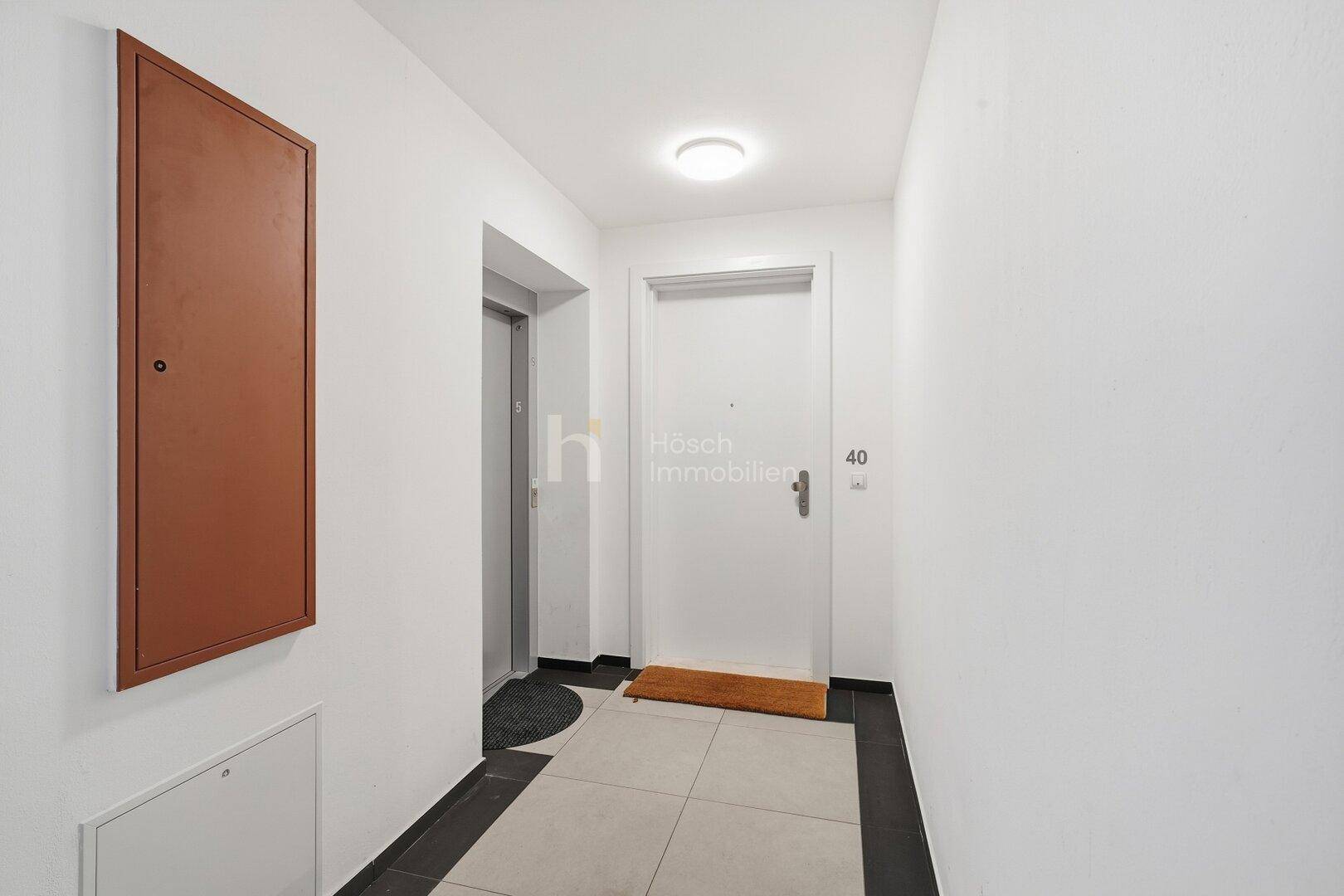 Moderner Flur mit hellem Fliesenboden, weißer Tür und Zugang zum Aufzug.
