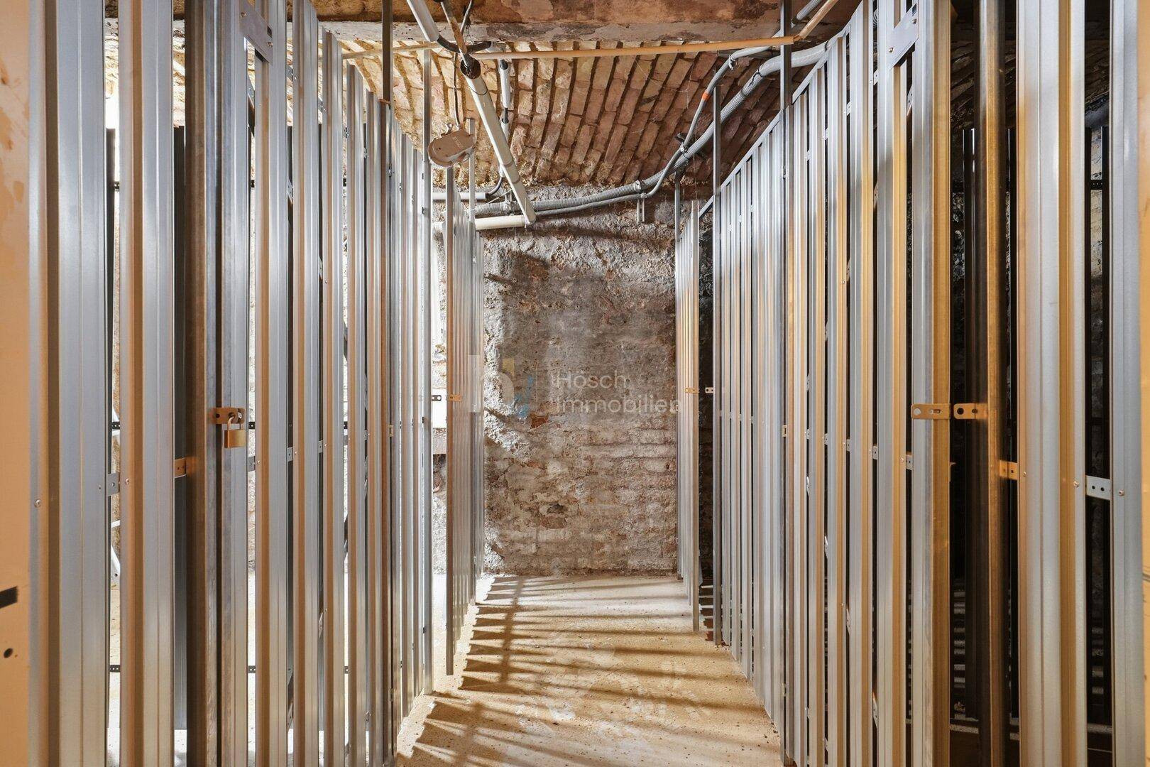 Kellerraum im Rohbau mit Metallständerwerk und sichtbaren Ziegelwänden und Leitungen.
