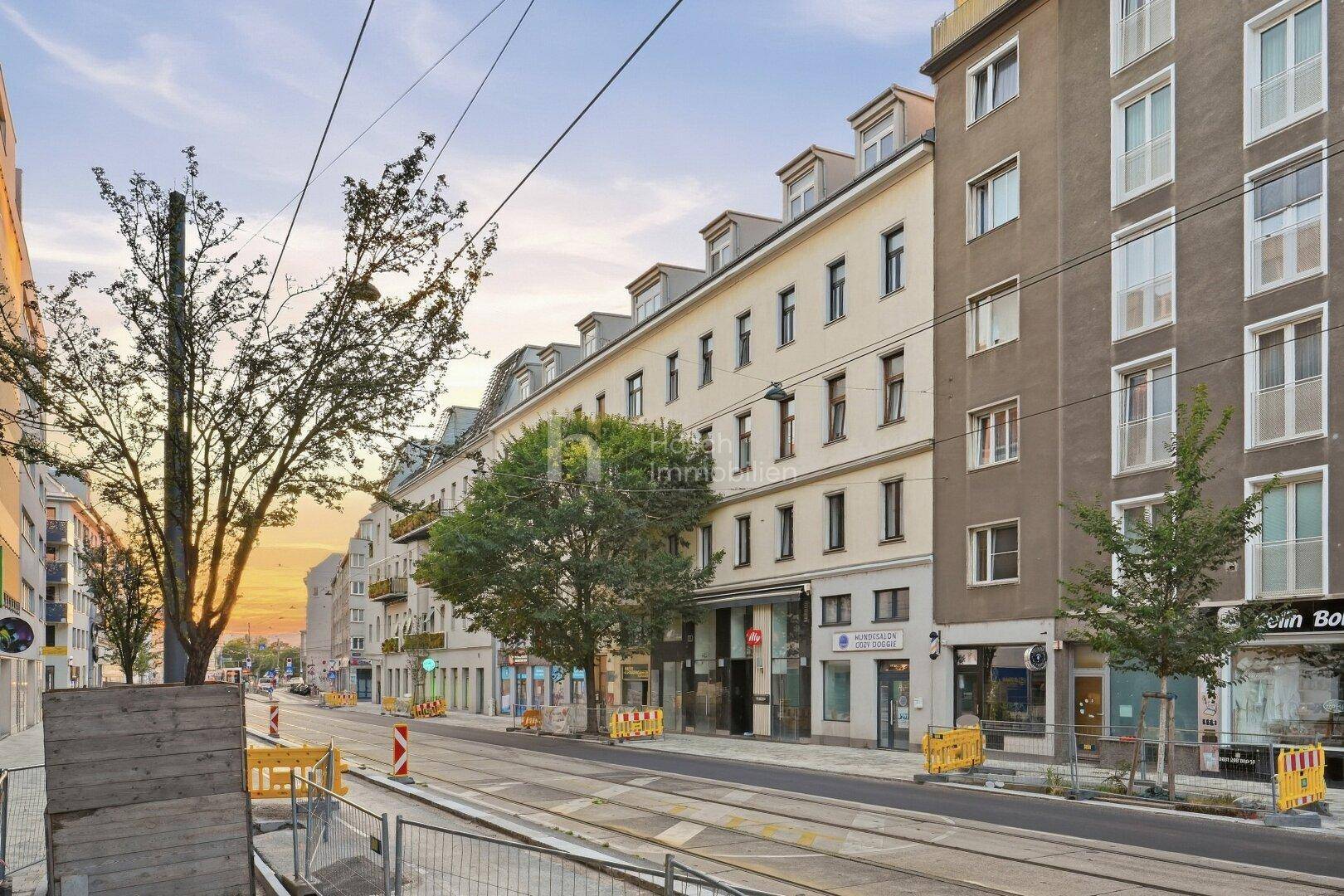 Gebäudefassade bei Sonnenuntergang mit Geschäften im Erdgeschoss und Straßenbahnschienen im Vordergrund.