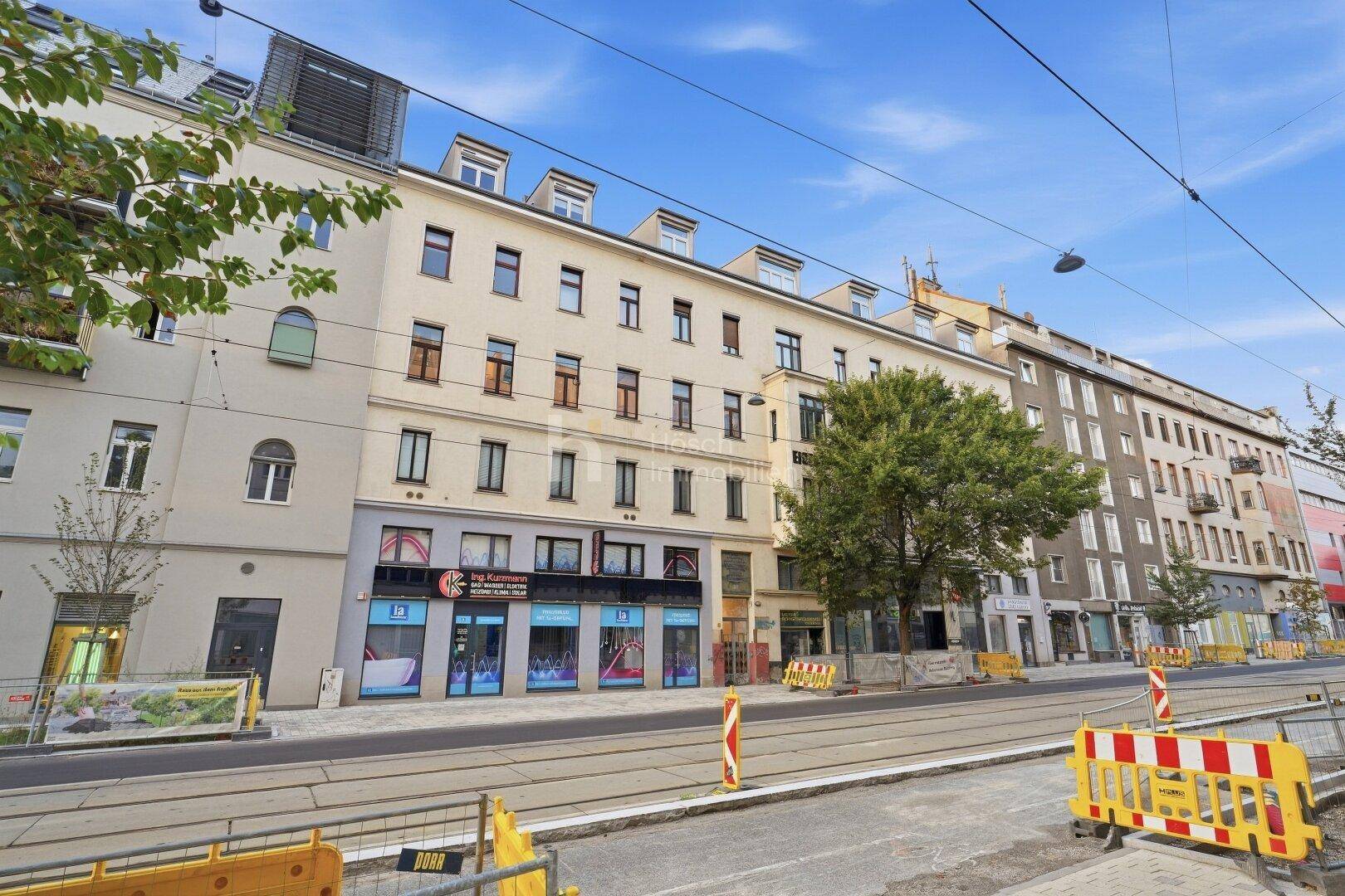 Gebäudefassade mit Geschäften im Erdgeschoss und Straßenbahnschienen im Vordergrund unter blauem Himmel.