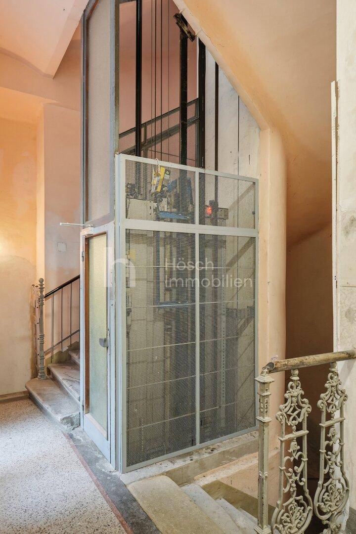 Treppenhaus mit offenem Aufzugsschacht und Marmorstufen, die den historischen Charakter betonen.