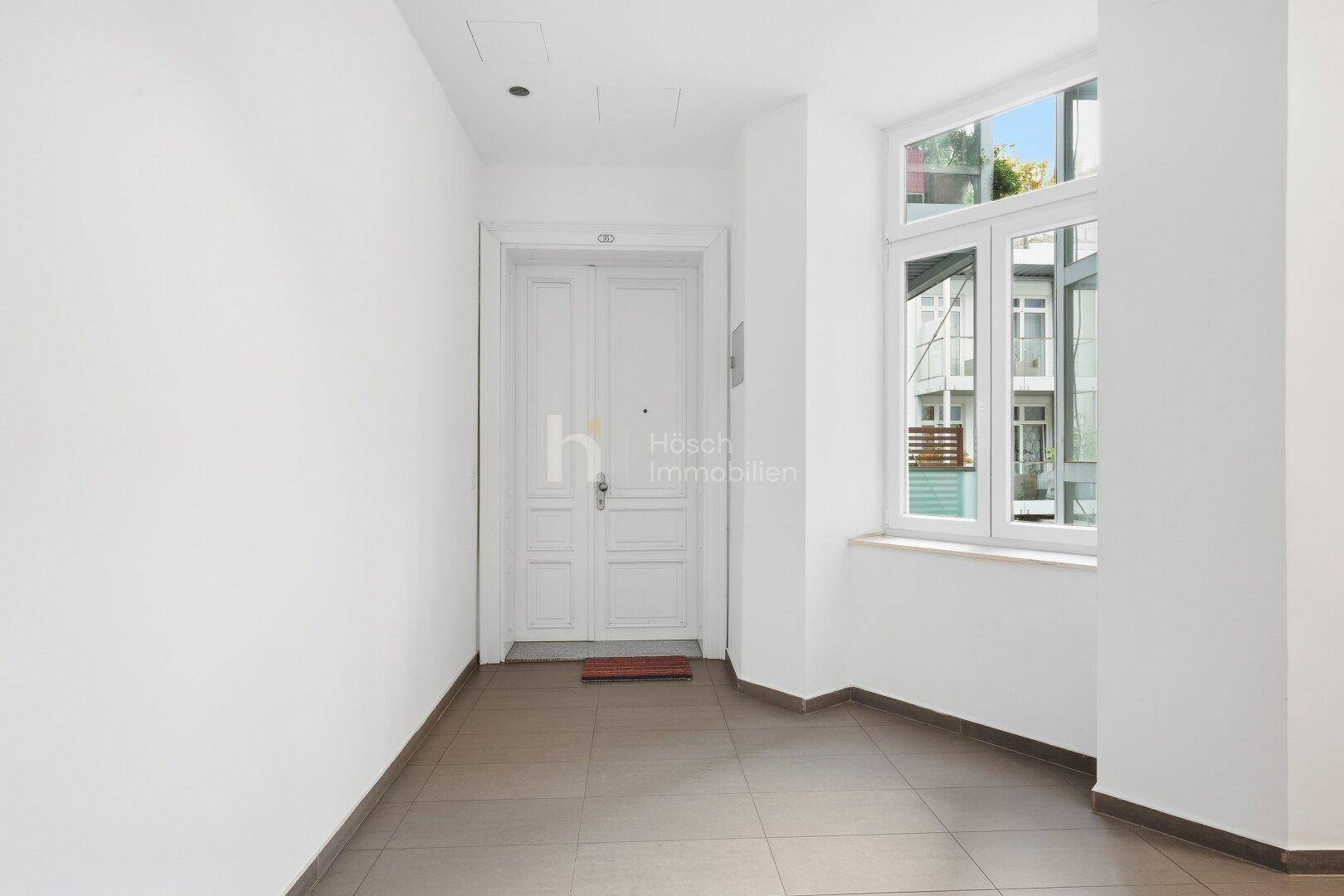 Heller Flur mit großem Fenster, das Tageslicht hereinlässt, und einer klassischen Wohnungstür.